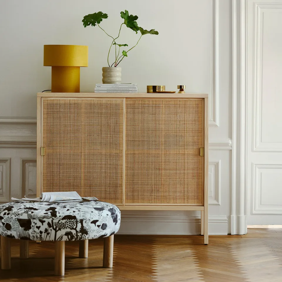 Modern living room with light wood cabinet, yellow lamp, plant vase, books, and floral pouffe.