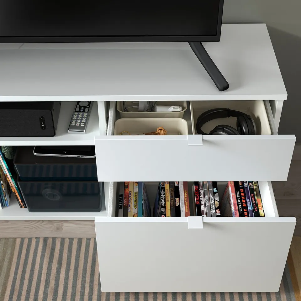 A white VIHALS tv bench with two drawers, one open displaying books, and another with headphones. The top holds a flat-screen tv.