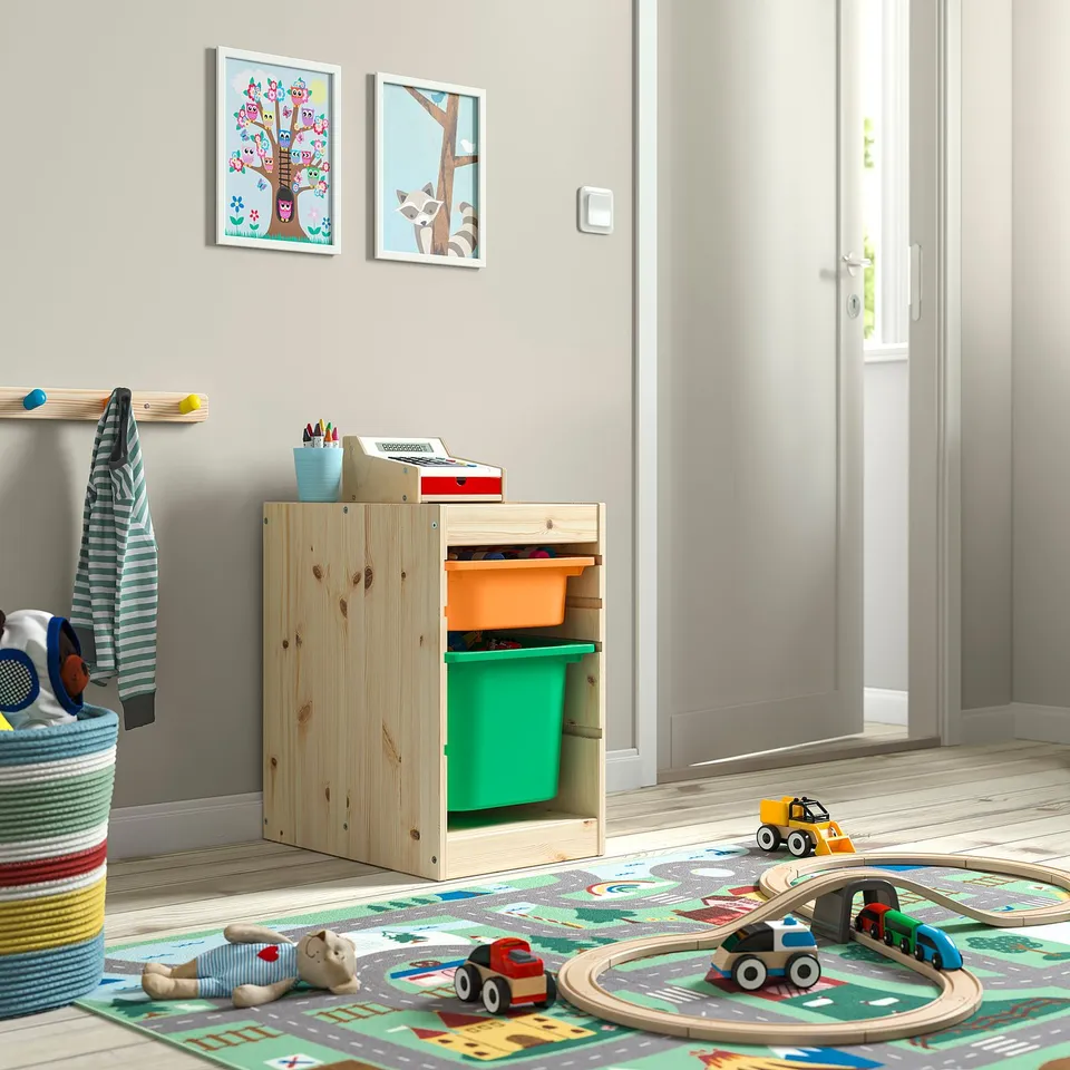 Wooden TROFAST kids storage unit with green & orange bins sits beside a colourful toy mat with a train track, toys, and a door.