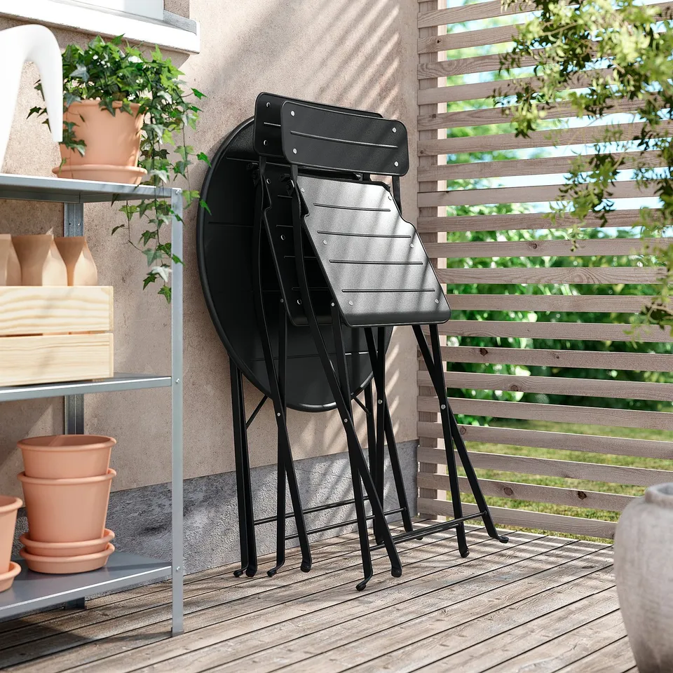Stack of 3 black metal SUNDSÖ folding chairs on balcony. Wooden fence, plants, and shelf with pots in background.