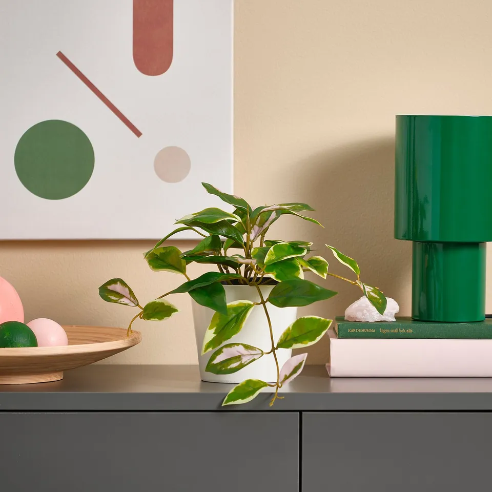 Small white potted artificial plant with green leaves sits on a table, surrounded by books and a green lamp.