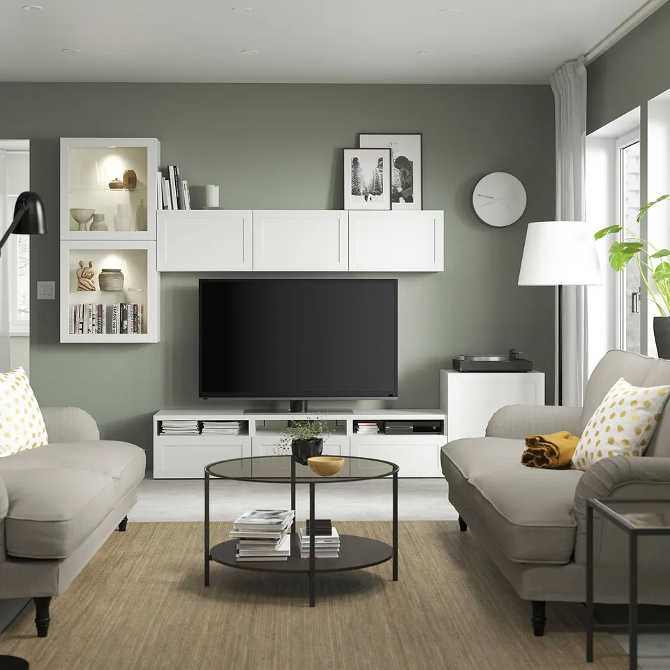 Modern living room with sage green walls, tv, grey sofas, dark coffee table, white storage, lamps, clock, pictures, pillows, and a plant.