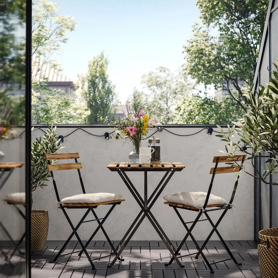 Small balcony with a TÄRNÖ table and chairs, light wood and black metal, decorated with string lights and flowers.