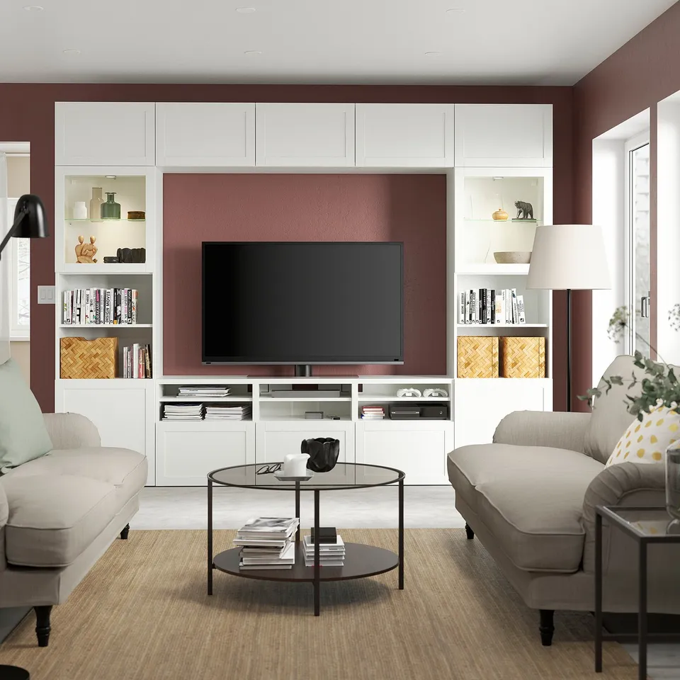 Modern living room with BESTÅ tv unit, flat-screen tv, books, and decorative items. Beige sofas, glass coffee table, and burgundy walls.