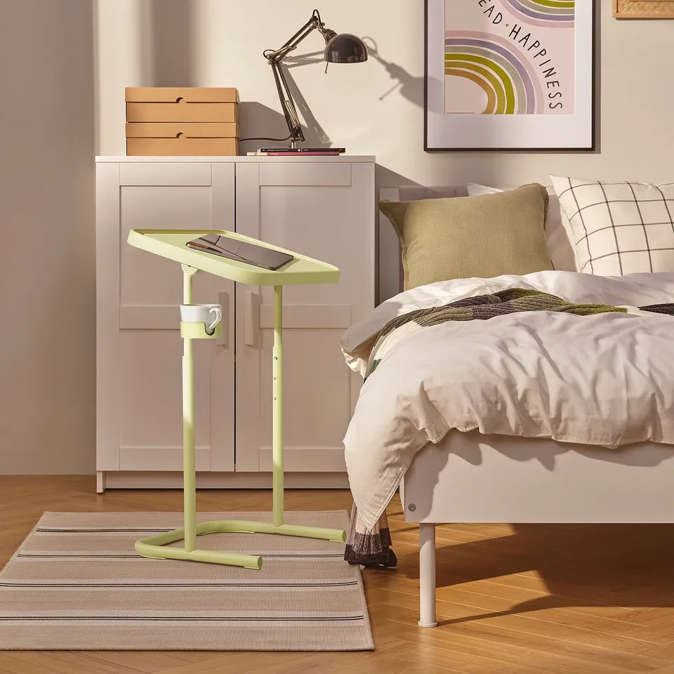 Small yellow-green BJÖRKÅSEN laptop stand next to a large bed, white cupboard, and a striped rug.