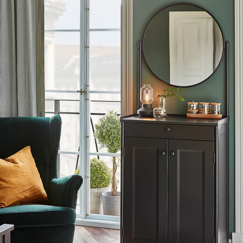 A cosy bedroom with a green armchair, a black tärnbry lamp, and a wooden cabinet with a round mirror.