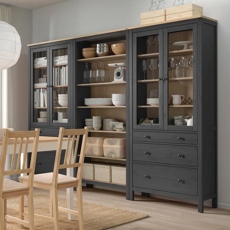 Modern dark grey HEMNES storage with glass doors, wooden accents, and open shelves for dishes and glassware, standing tall beside dining chairs.