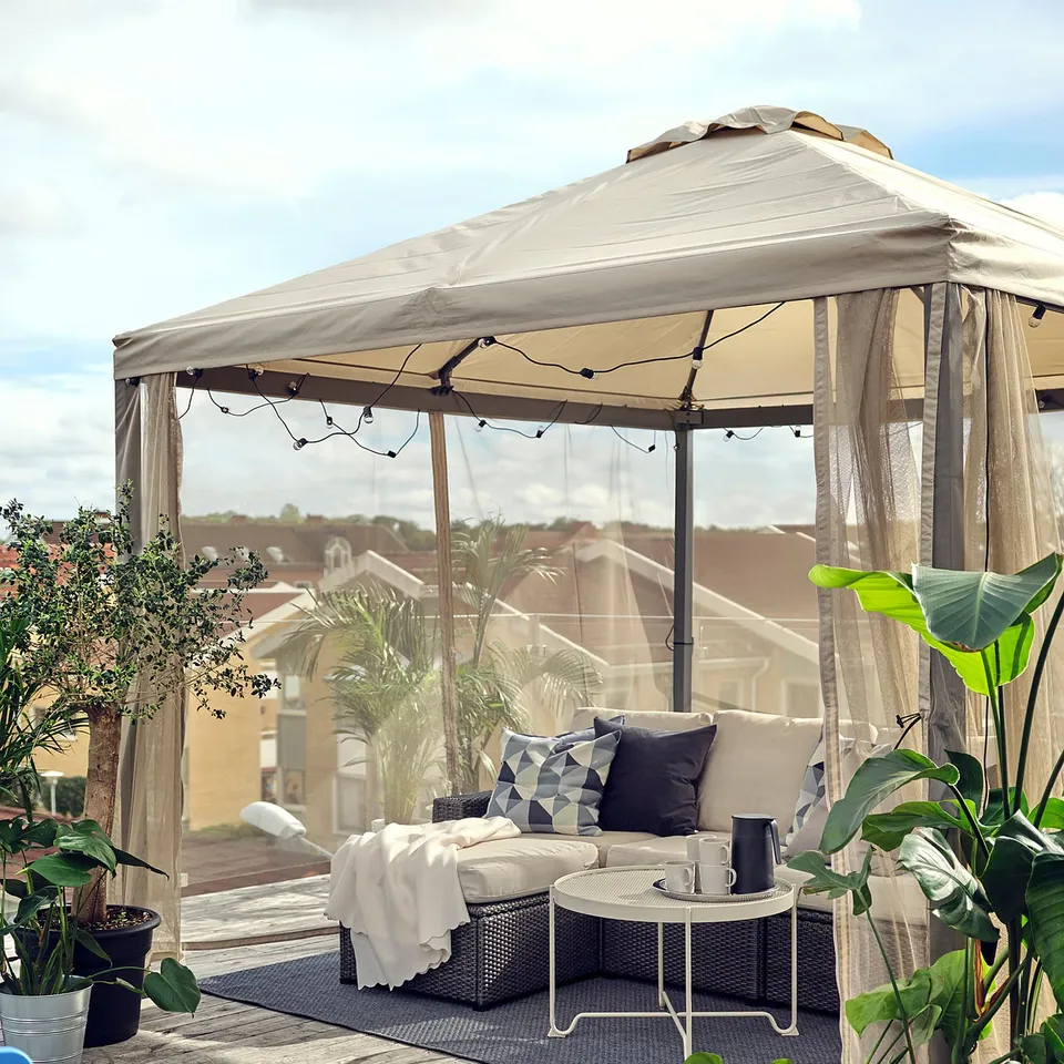 Cosy outdoor gazebo with cream-colored canopy, wicker sofa, striped cushions, and potted plants.