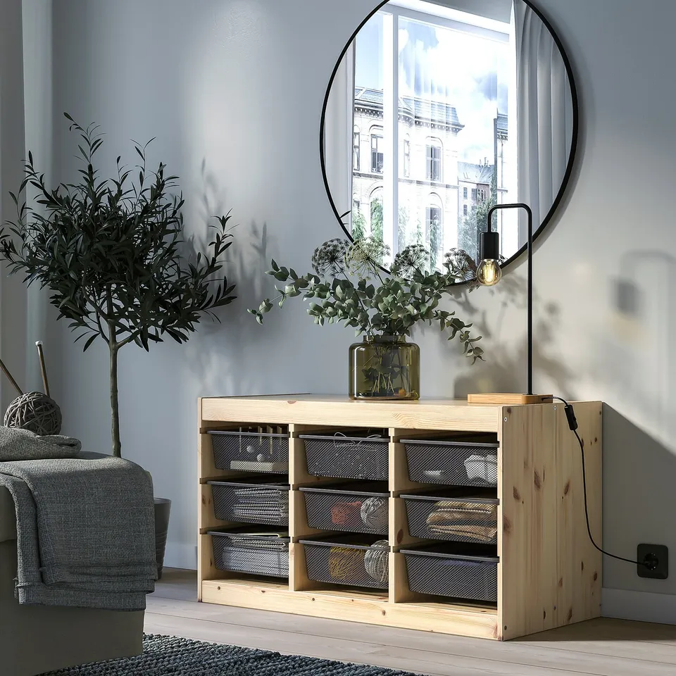 The TROFAST storage unit, made of pine wood with mesh boxes, sits beneath a large round mirror and beside a tall indoor plant in a bright room.