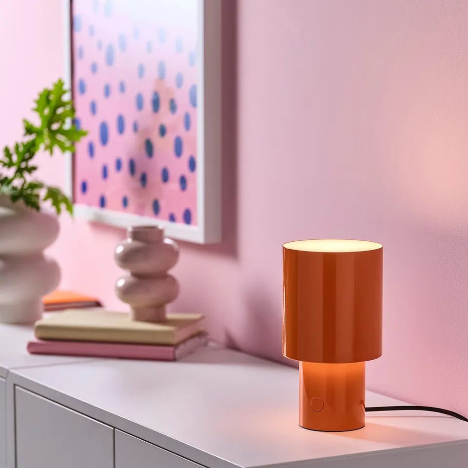 Orange SPETSBOJ lamp on white surface, soft glow, with books, plant, pink decor.