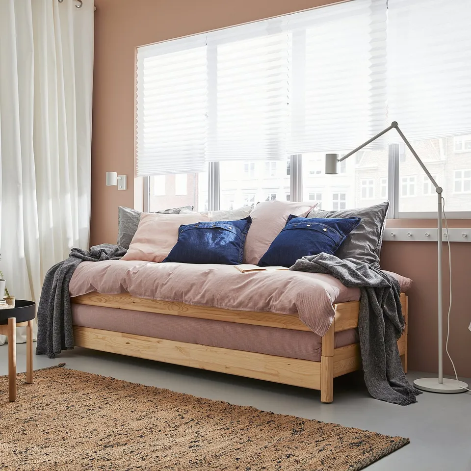 Modern cosy bedroom with a UTÅKER wooden bed, light pink bedding, blue pillows, grey throws, wooden bedside table, and floor lamp by window.