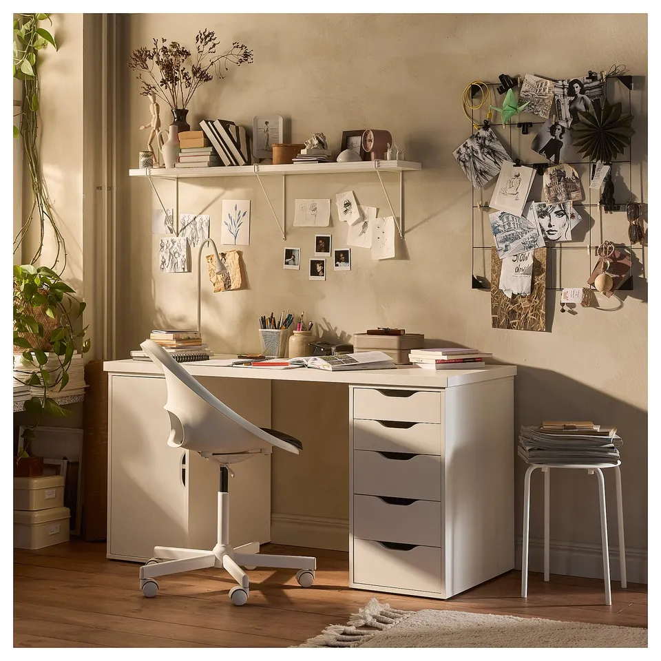 Modern home office with white LAGKAPTEN desk featuring drawers and a door. Decorated with plants, books, and art on a beige wall.