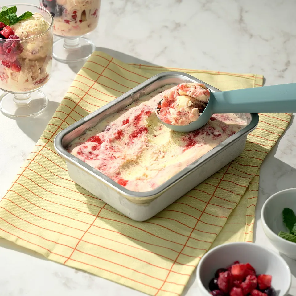 Pink berry ice cream in container with scoop, glasses, bowl on yellow cloth.