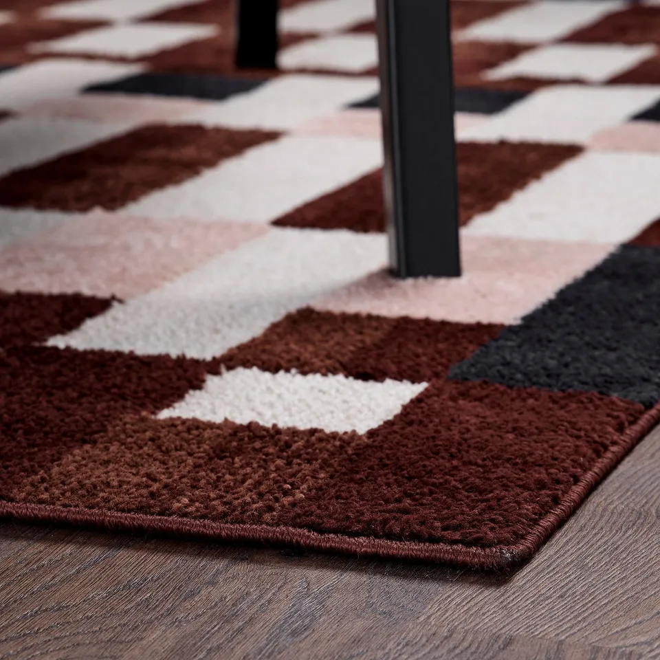 A patterned rug with red, pink, and beige checks. Placed near a black furniture leg. Soft, low-pile for comfort.