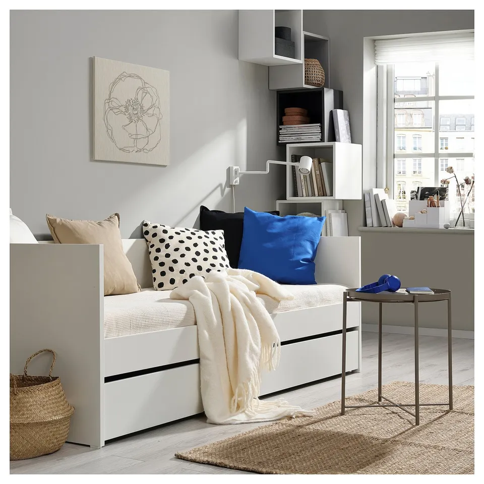 Cosy modern living space with white daybed, colourful pillows, and built-in shelves. Large city window; minimalist design.