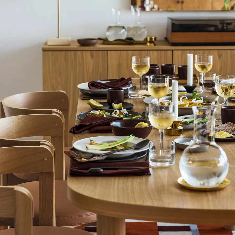 Elegant dining setup with wooden chairs, oval table, black plates, glasses, wine, food, and candles. Sparse decoration.