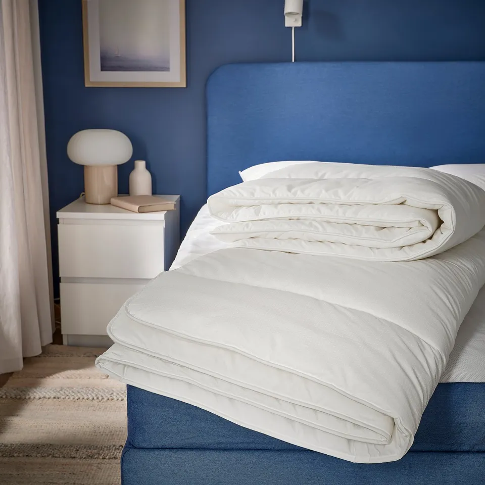 Bedroom with blue SKOGSFRÄKEN bedding; white lamp, books on small table beside large bed; blue headboard, wall art.