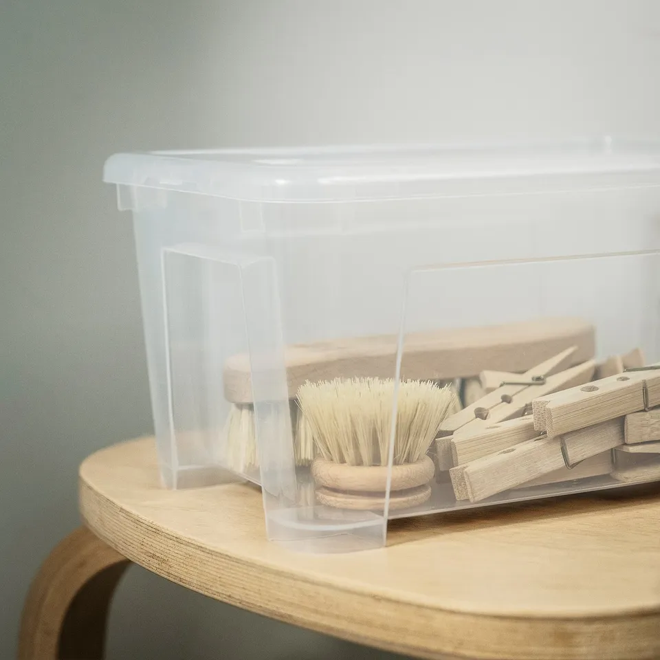 Clear plastic storage box with wooden clothespins and brushes, placed on a wooden surface.