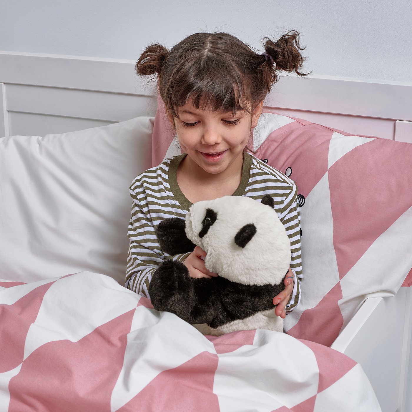 A young girl in striped pyjamas sits in bed, hugging a soft panda toy, symbolising comfort and companionship.