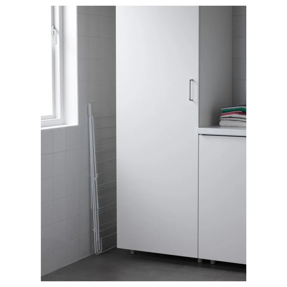 Metal drying rack leans against wall, beside a large white cabinet. Perfect for drying clothes indoors or outdoors.