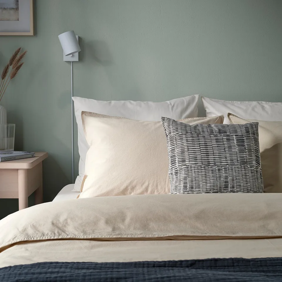 Bed with white pillows, one patterned. Green wall, bedside lamp and books.