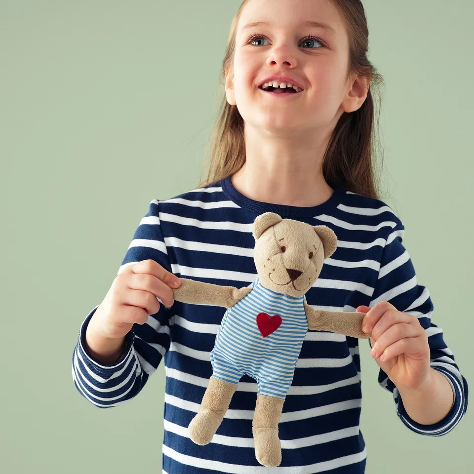 A young girl holds a plush teddy bear wearing a blue striped shirt with a red heart, both smiling happily.
