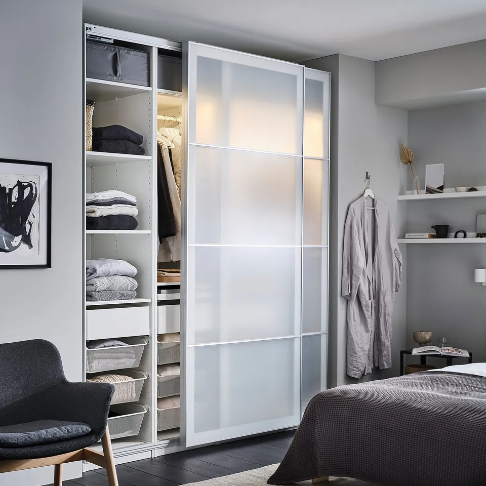 Modern bedroom with white PAX wardrobe, glass doors, grey chair, hanging robe, decorative items, and dark bed with folded blanket.