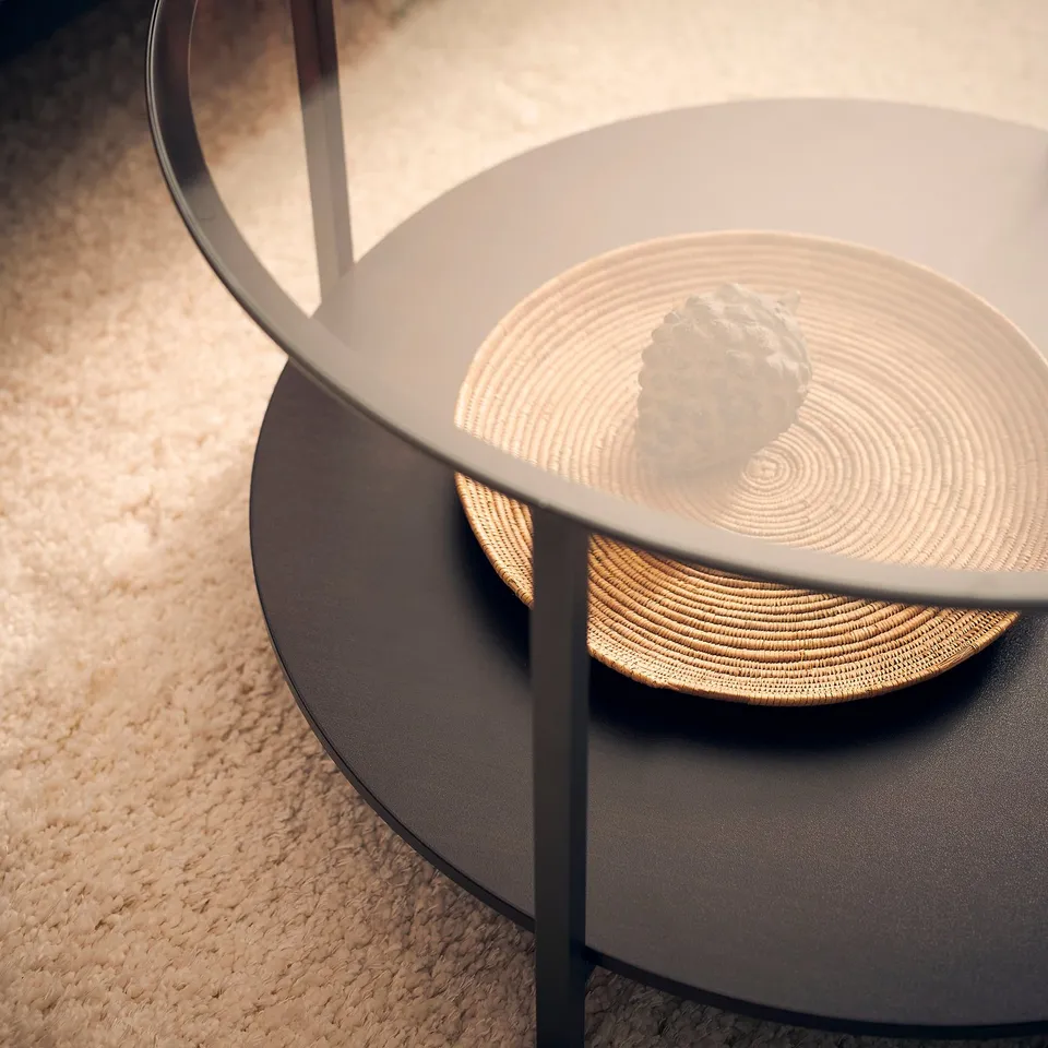 Modern, round coffee table VITTSJÖ with glass top and black shelf, displaying a woven tray and a decorative rock.