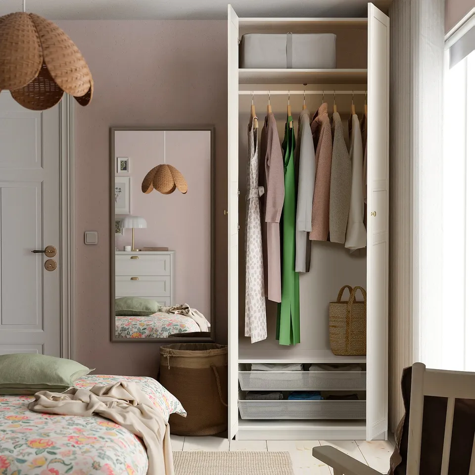 Organised bedroom with PAX wardrobe, hanging clothes, shelves, mesh baskets, pink wall, mirror, patterned bed.