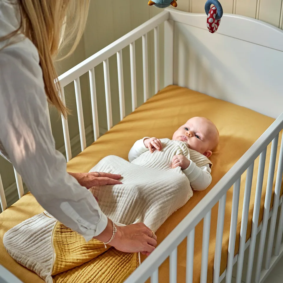 Blonde person adjusting sleep bag on baby in white crib, ensuring comfort with soft organic cotton.