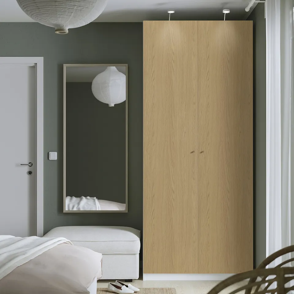 A modern bedroom a tall, light-colored wood PAX wardrobe beside a large mirror and a white bed.