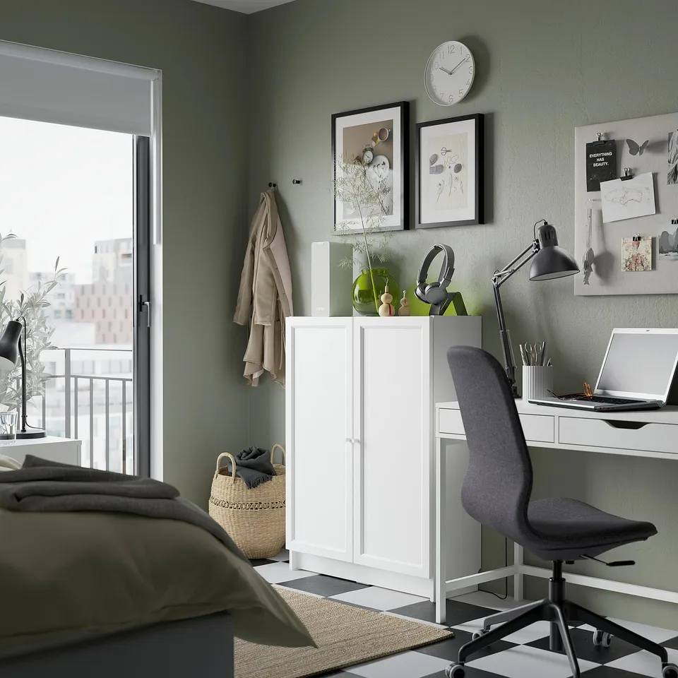 Cosy bedroom with green walls, featuring a white BILLY bookshelf, grey desk, and office chair. A clock, framed art, and plants decorate the wall.