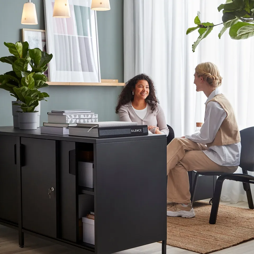 Two people chat by a TROTTEN room divider cabinet with books and a plant on top.