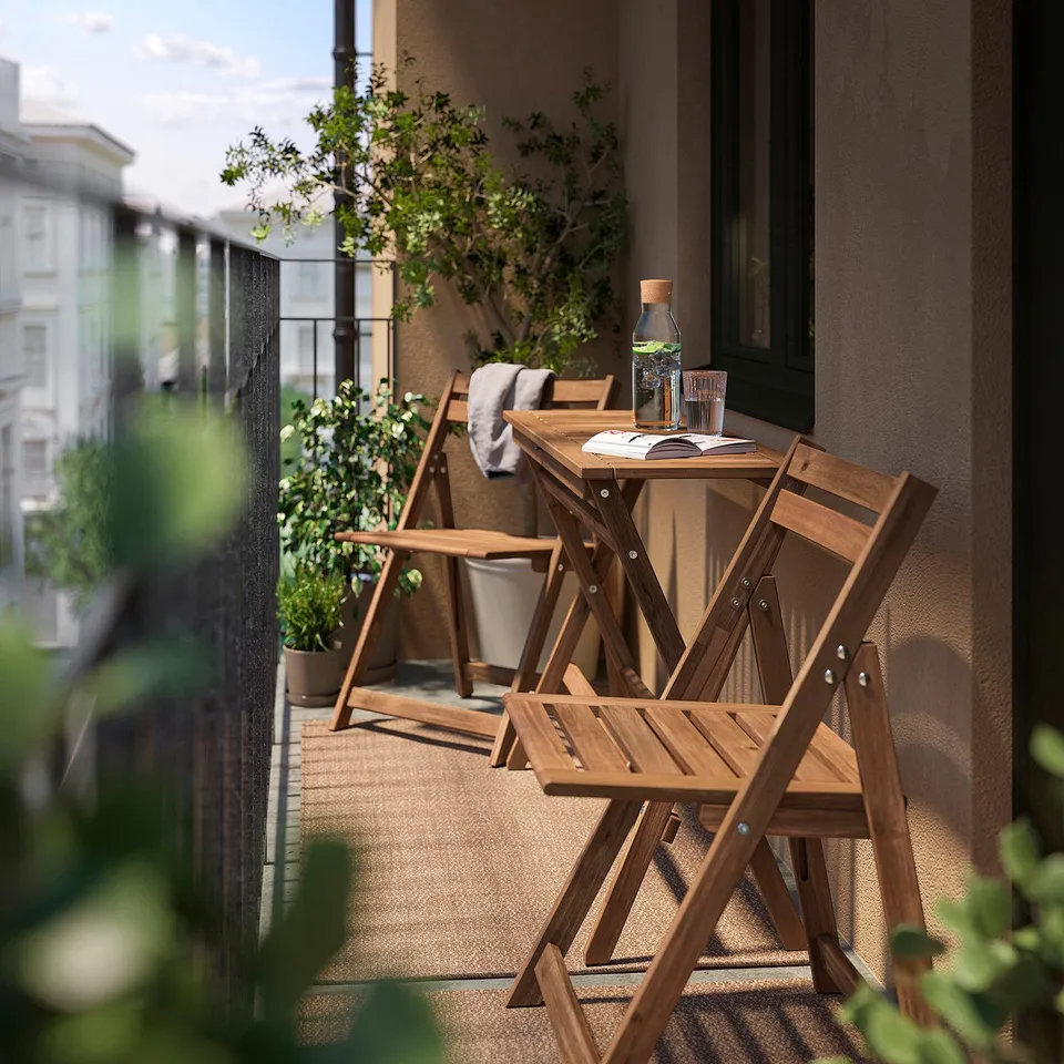 Small balcony with brown NÄMMARÖ wooden table and chairs, featuring a glass carafe, cups, and a magazine.