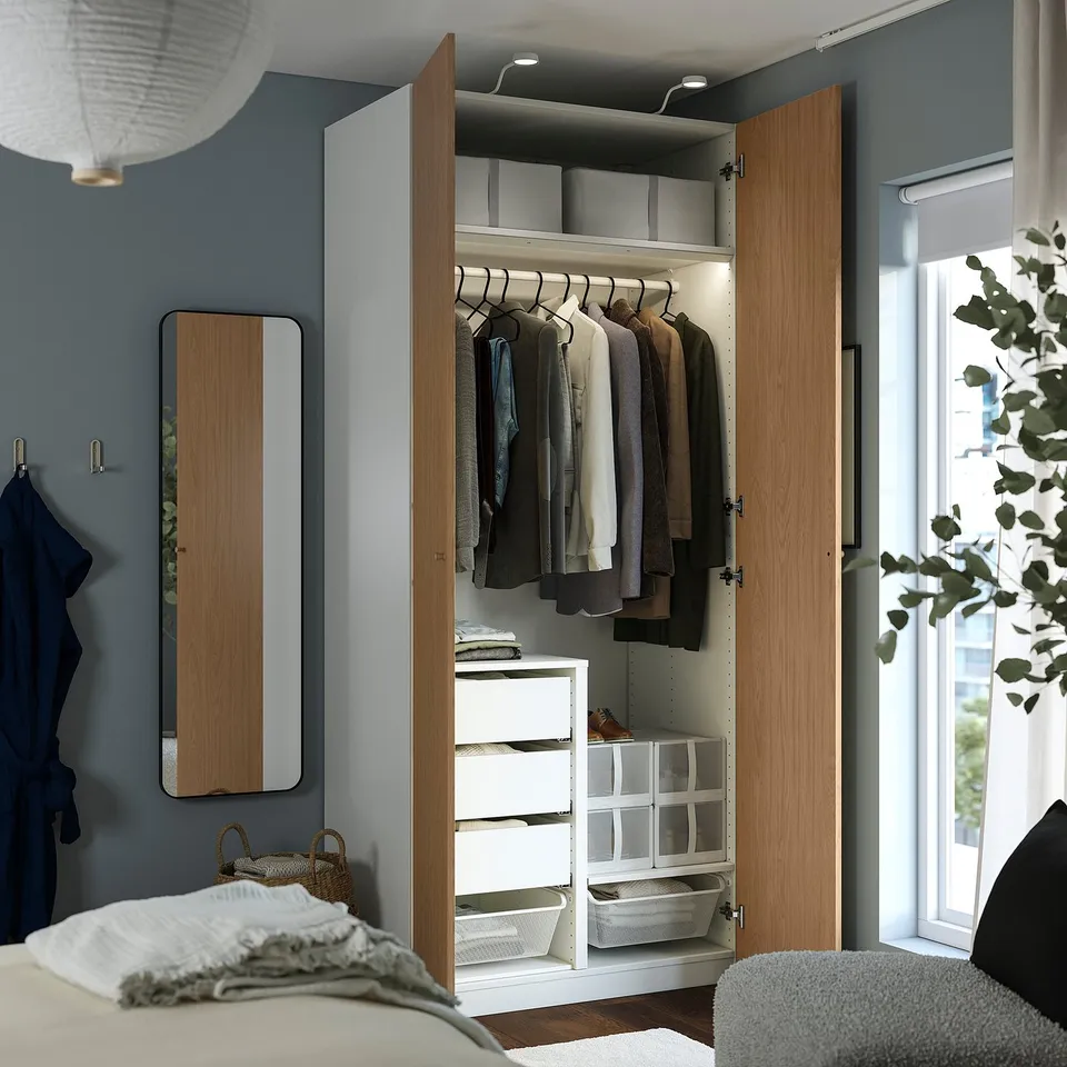 A modern wooden wardrobe named PAX stands open, displaying internal storage spaces including hangers with clothes, drawers, and shelves with baskets.