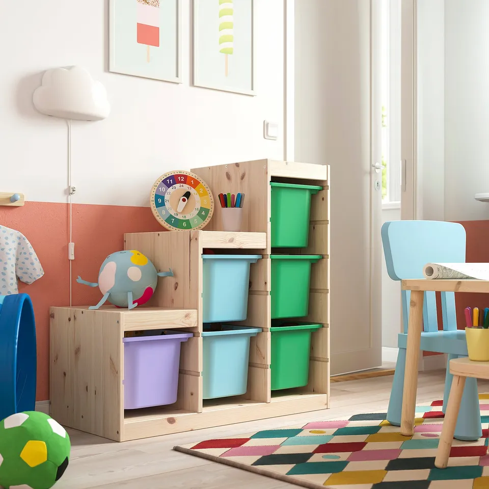 A TROFAST storage unit in a childs room with colourful boxes, a cuddly toy, wall clock, blue chairs, and a colourful rug.