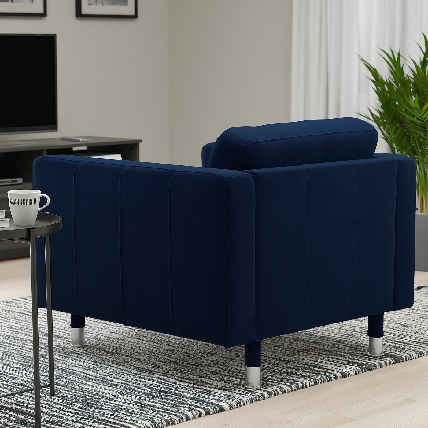 A modern living room with a navy blue armchair, a small side table with a coffee mug, and a tv on a wooden stand. A plant and a rug are also present.