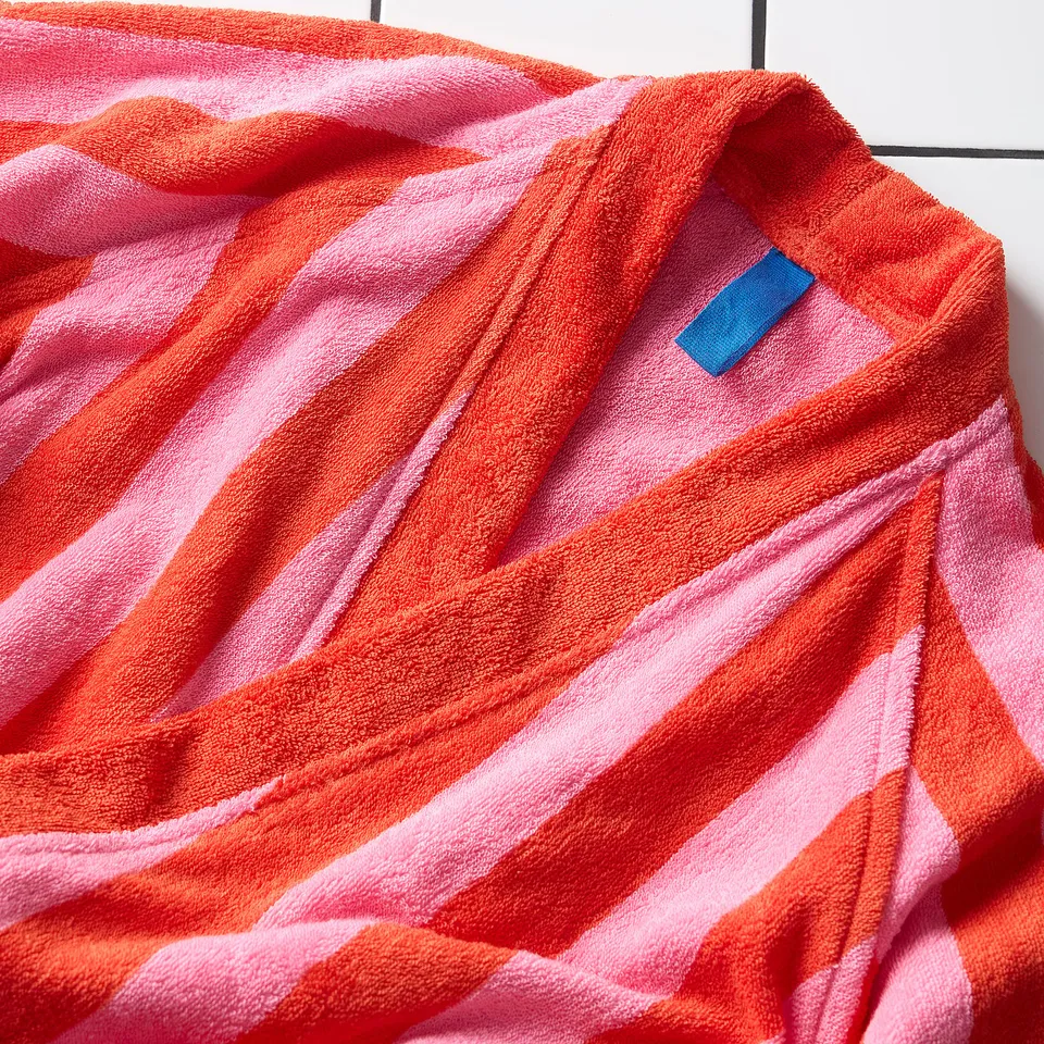 Vibrant, striped bathrobe with bright red and pink hues, featuring a soft, absorbent cotton terry texture. A blue tag is visible inside the collar.