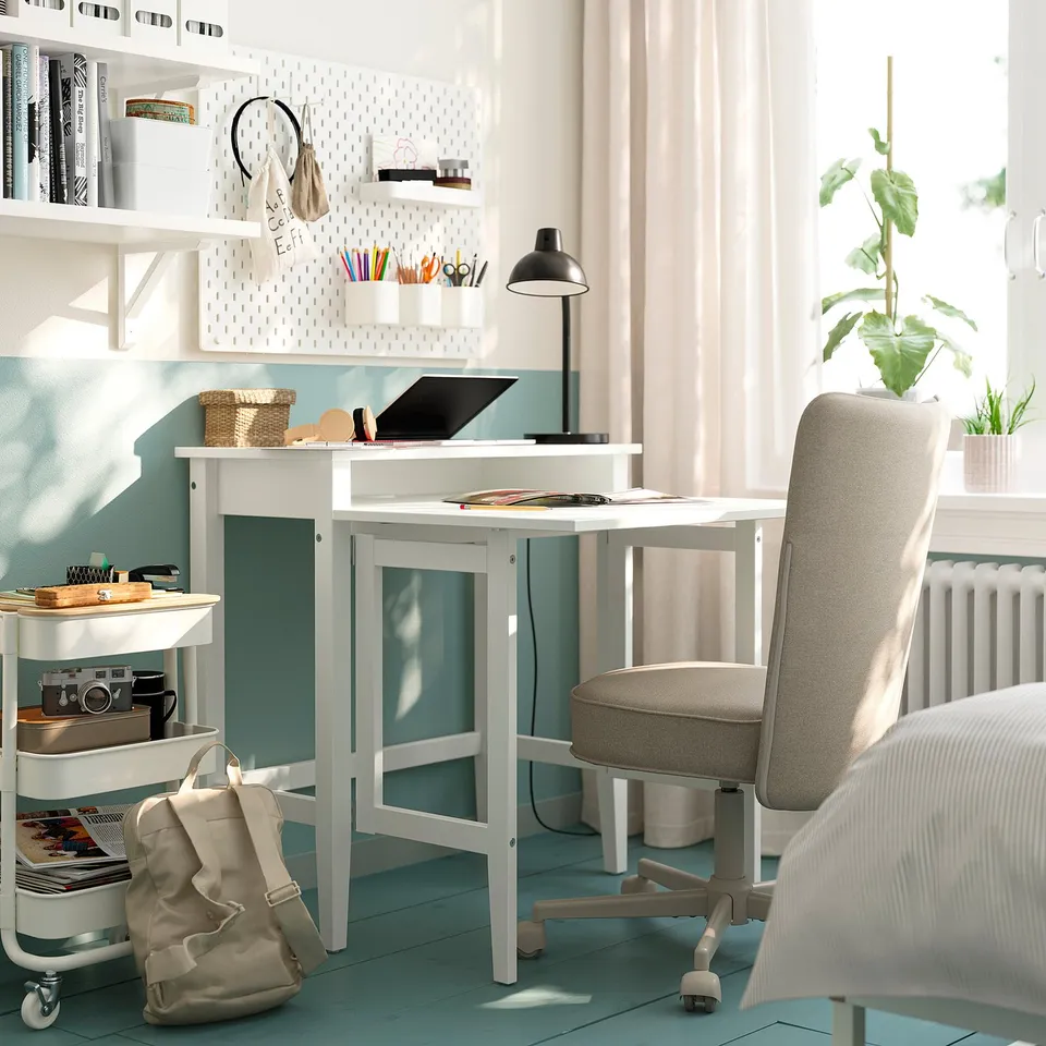 Cosy home office setup with white GULLABERG desk and grey chair. Desk holds laptop, lamp, and accessories; surrounded by bookshelves and plants.