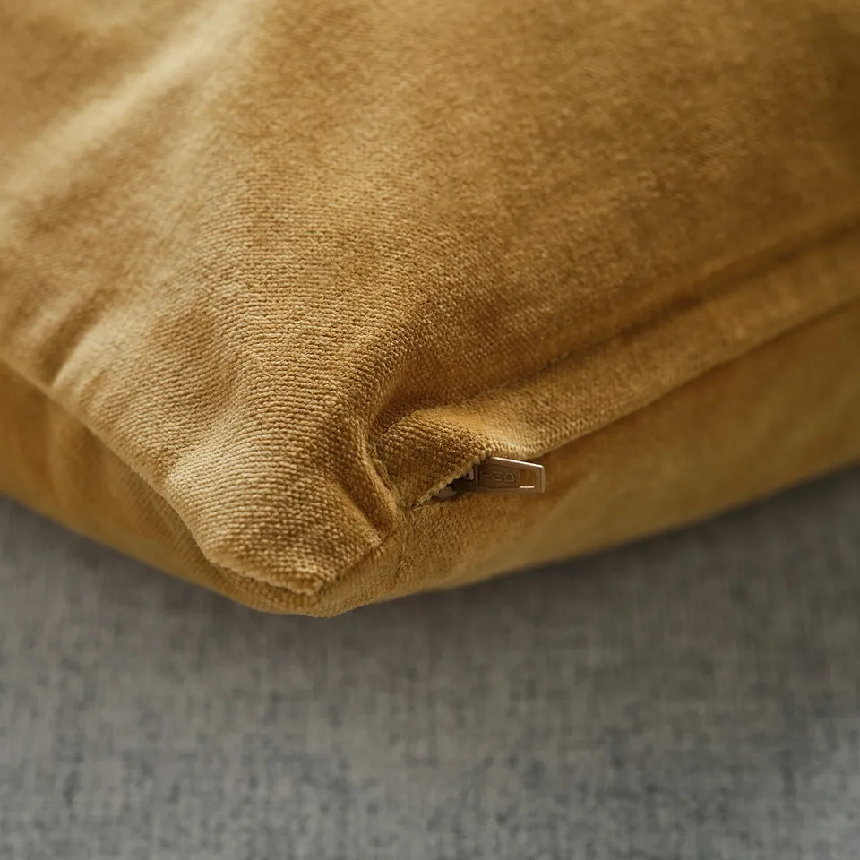 Close-up of a velvety, mustard-colored cushion cover with a subtle sheen. Hidden zip visible, resting on a textured surface.