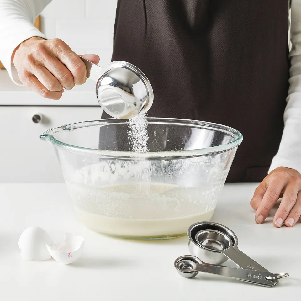 A person pours sugar from a measuring spoon into a bowl; measuring spoons used.