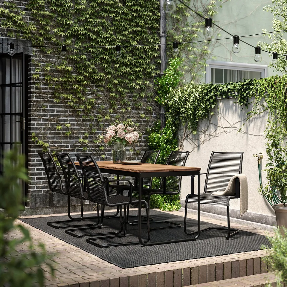 Modern outdoor dining set with extendable table and six black chairs. Surrounded by greenery and string lights.