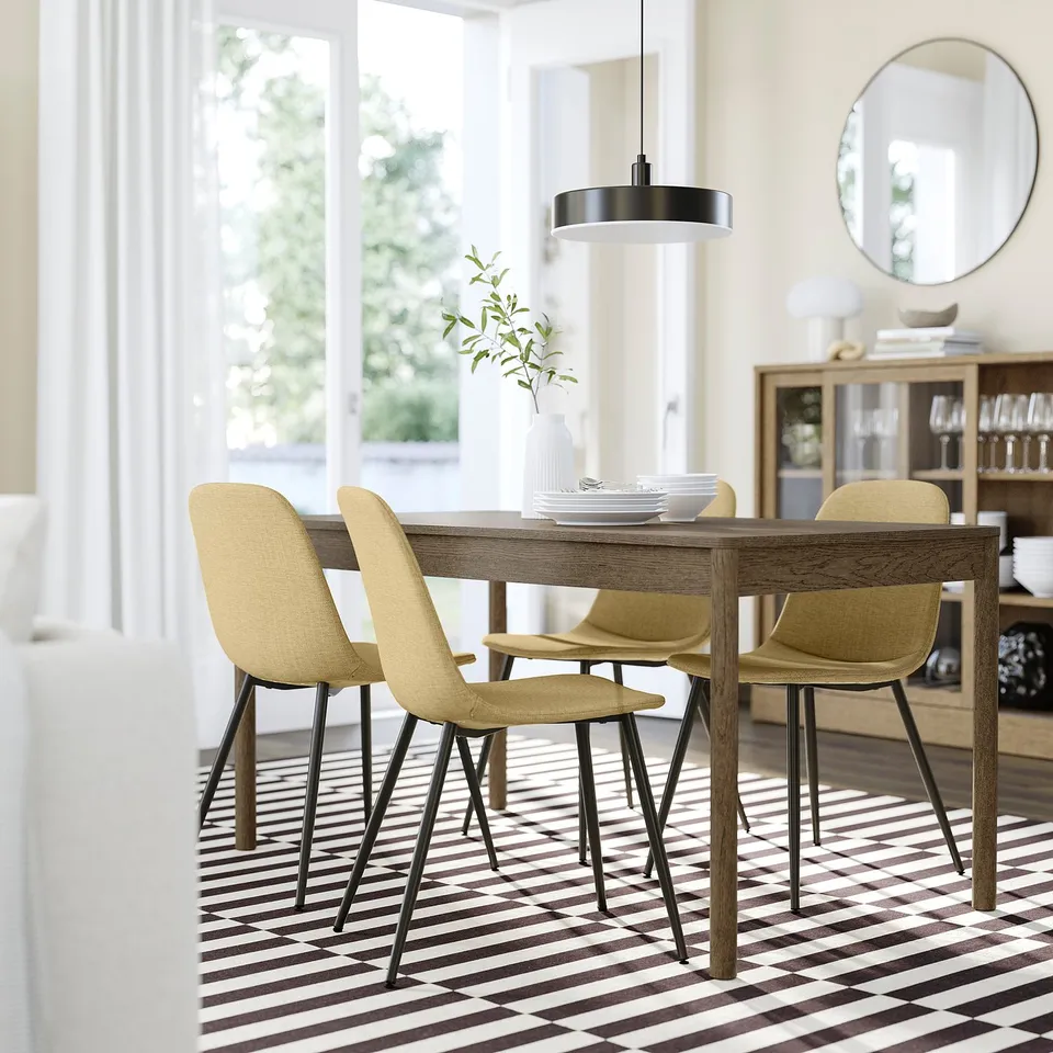 Modern dining area with rectangular table, four beige chairs, patterned tiles, and hanging lamp. Sideboard with glassware and large window.