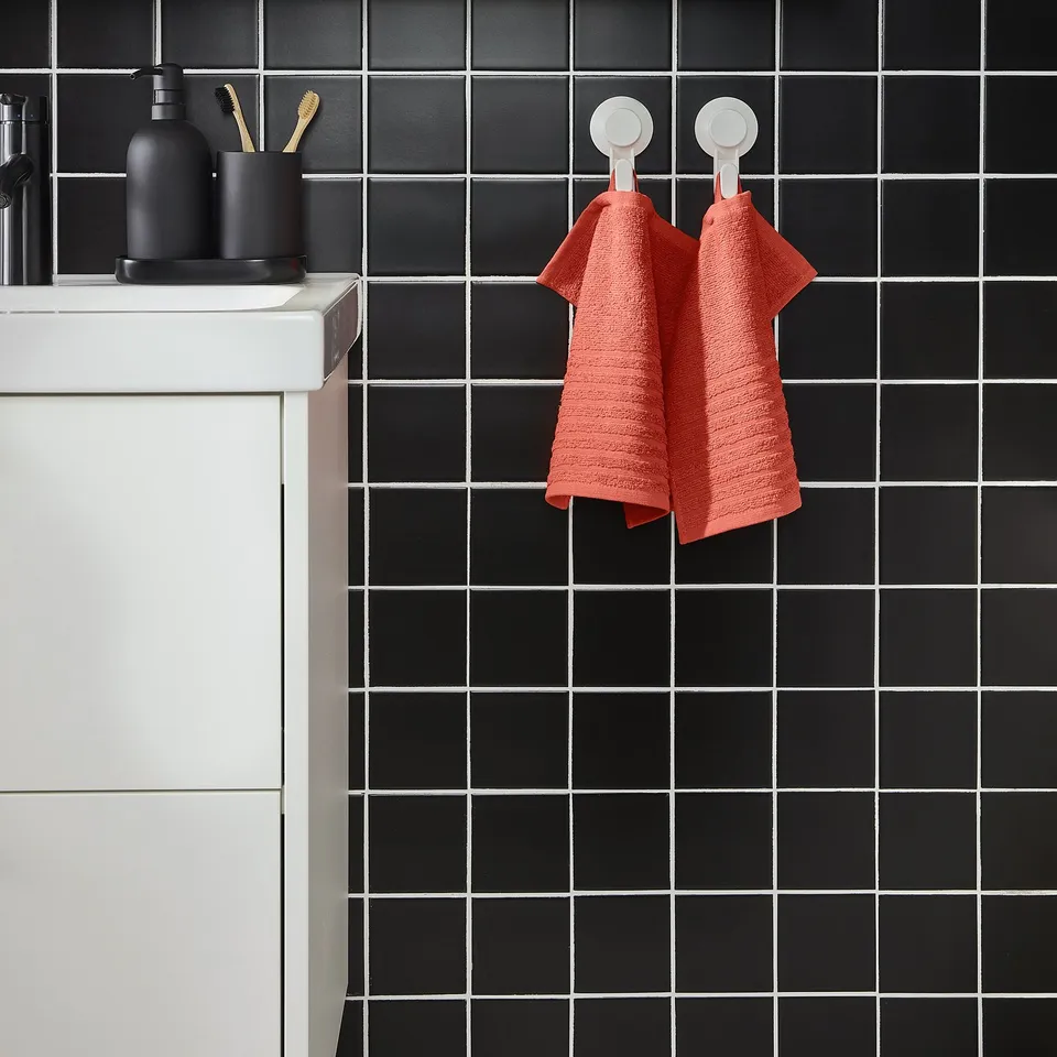 Modern bathroom with black tiled wall, two white suction cup hooks hanging an orange VÅGSJÖN towel, smaller than adjacent sink and black accessories.