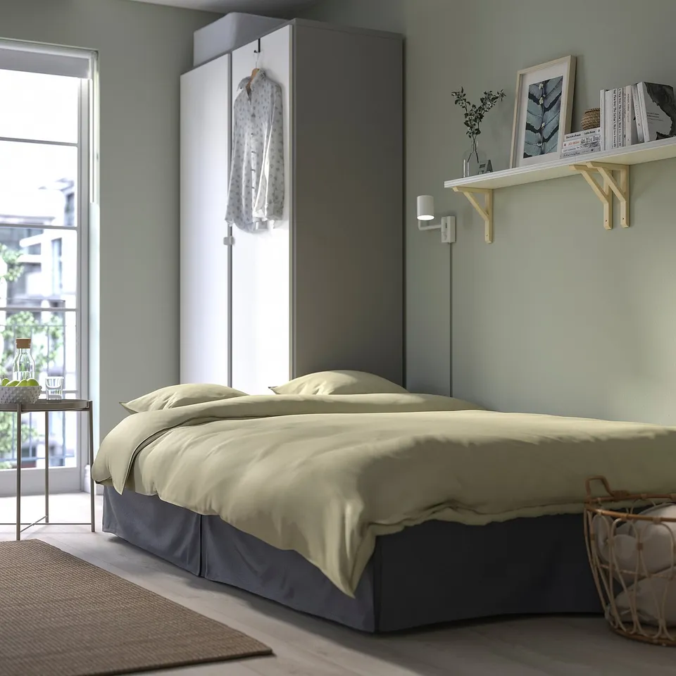 Cosy bedroom with beige bed, white shelves, green walls. Bed has grey skirt, hanging robe, wall light, and basket beside bed.
