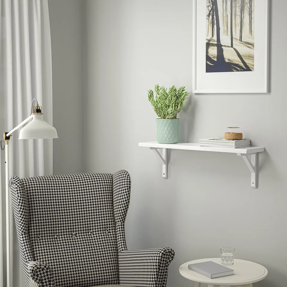 Cosy reading nook with TRANHULT white wall shelf displaying books and plants. Houndstooth armchair beside, with white lamp and framed nature photo.