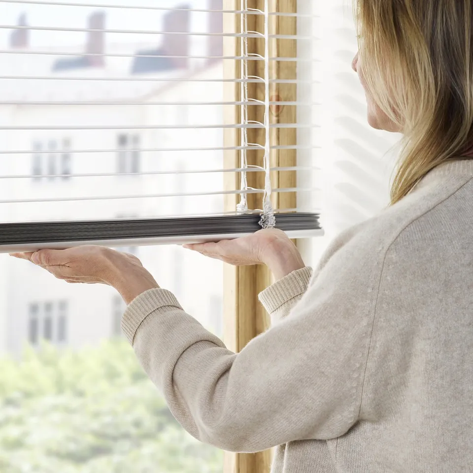 A person adjusts modern cordless venetian blinds near a window. The person tilts the slats for light control and privacy.