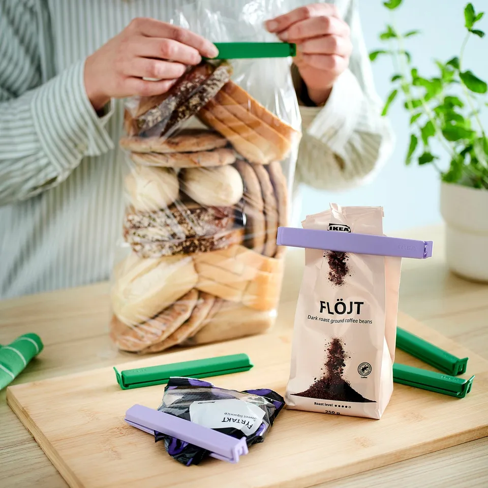 A person secures a bread bag with green BEVARA clips; a coffee bag and more clips sit on a wooden table.