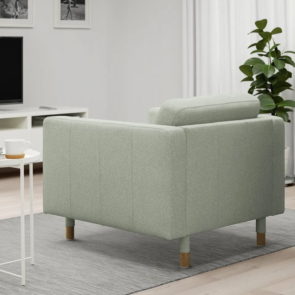 Modern light green armchair with wooden legs and cushions, near plant and coffee cup.
