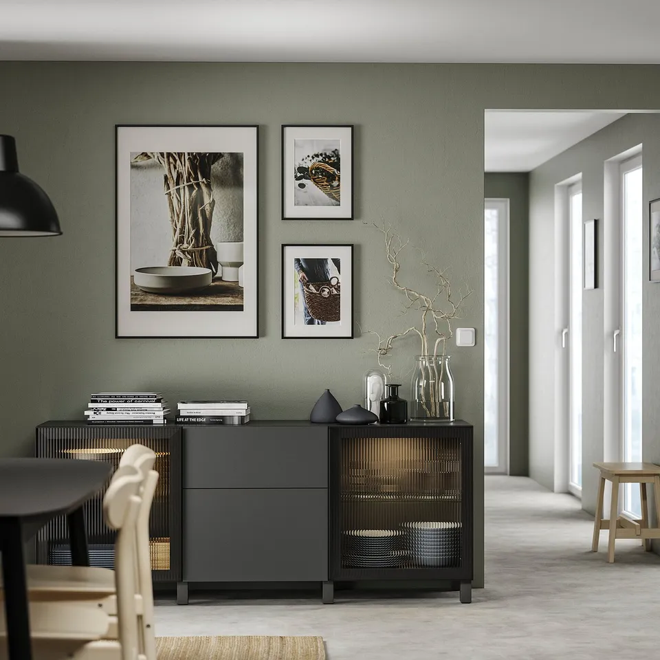 Contemporary living room with sage green walls, minimalist black sideboard, white framed art, and light wood chair.